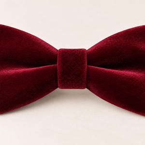 May include: A deep burgundy red bow tie crafted from a soft velvet fabric. The bow tie features a classic double-layered shape with a central knot detail, showcasing a smooth, light-reflecting texture across the entire surface of the accessory.