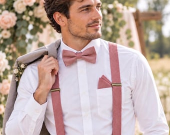 May include: A crisp white button-down shirt paired with dusty rose suspenders, a matching dusty rose bow tie, and a coordinating dusty rose pocket square. A light gray suit jacket is draped over one shoulder, completing the formal ensemble.