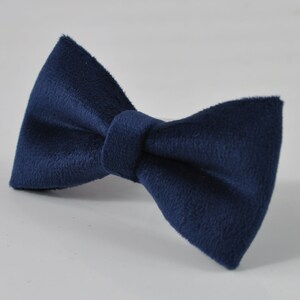 May include: A navy blue bow tie made of a soft, textured fabric. The bow tie is a classic accessory, perfect for formal events or adding a touch of sophistication to any outfit. The bow tie is centered on a white surface.