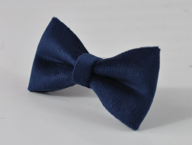 May include: A navy blue bow tie made of a soft, textured fabric. The bow tie is a classic accessory, perfect for formal events or adding a touch of sophistication to any outfit. The bow tie is centered on a white surface.