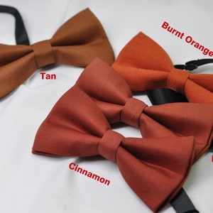 May include: Four bow ties in different shades of brown: tan, burnt orange, cinnamon, and rust. The bow ties are arranged in a row on a white surface.