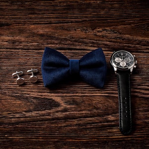 May include: A navy blue velvet bow tie, silver cufflinks, and a black leather strap watch with a silver case are arranged on a dark wood surface. The watch face has multiple dials and hands.