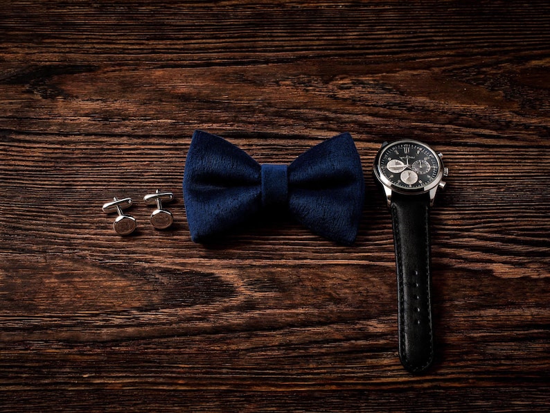 May include: A navy blue velvet bow tie, silver cufflinks, and a black leather strap watch with a silver case are arranged on a dark wood surface. The watch face has multiple dials and hands.