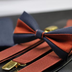 May include: A set of brown braces with gold metal clips and a matching bow tie with navy blue and rust orange fabric.