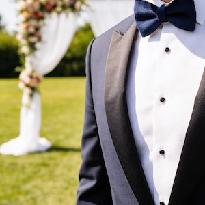 May include: A navy blue tuxedo jacket with black lapels and a white dress shirt. A navy blue bow tie is worn with the tuxedo. The background includes a floral wedding arch and green grass.