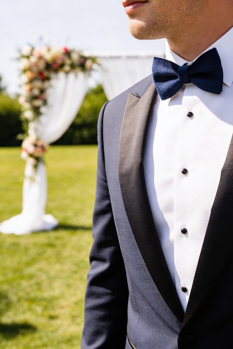 May include: A navy blue tuxedo jacket with black lapels and a white dress shirt. A navy blue bow tie is worn with the tuxedo. The background includes a floral wedding arch and green grass.