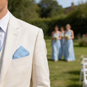 Puede incluir: Una chaqueta de traje color crema sobre una camisa blanca de cuello rígido y un pañuelo de bolsillo azul claro en el bolsillo del pecho. La chaqueta está confeccionada con solapa de muesca y un cierre de un solo botón frontal.