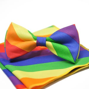 Può includere: Un papillon a righe arcobaleno e un fazzoletto da taschino abbinato. Il papillon è annodato e poggia sul fazzoletto da taschino piegato. I colori includono rosso, arancione, giallo, verde, blu e viola. Gli articoli sono in tessuto liscio e lucido.