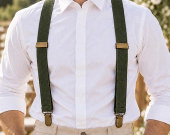 May include: A pair of olive green suspenders featuring brass-toned metal adjusters and clips, worn over a crisp white button-down shirt and light beige trousers. The suspenders have a textured fabric finish and are secured to the waistband.