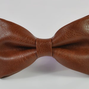 May include: A brown bow tie with a textured, leather-like finish. The bow tie features a classic pre-tied shape with a central knot detail, showcasing a consistent grain pattern across the surface of the material, presented against a plain background.