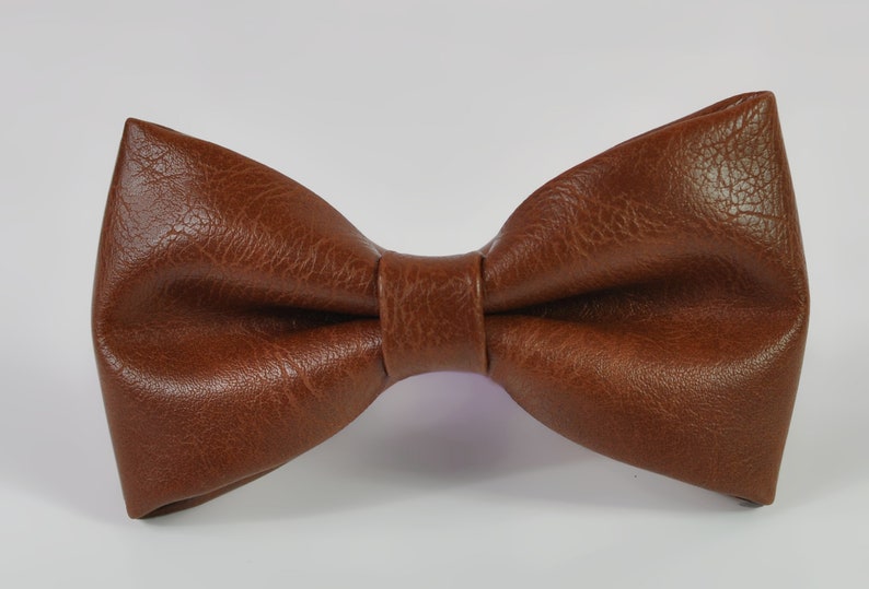 May include: A brown bow tie with a textured, leather-like finish. The bow tie features a classic pre-tied shape with a central knot detail, showcasing a consistent grain pattern across the surface of the material, presented against a plain background.