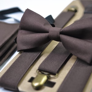 May include: A brown bow tie, suspenders, and a pocket square set. The bow tie is pre-tied and sits atop the suspenders, which have gold-colored metal clasps. The pocket square is folded neatly. The set is displayed on a cardboard backing.
