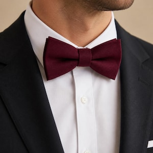 Puede incluir: Una pajarita de color burdeos intenso con un acabado de tela texturizada, combinada con una camisa blanca de botones y una americana negra. La pajarita presenta una forma clásica preanudada con un detalle de nudo central.