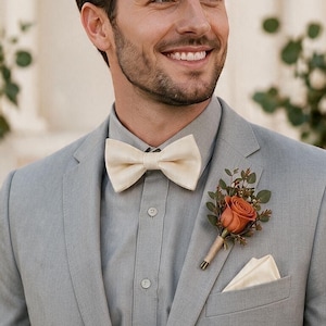 May include: A light gray suit jacket worn with a light gray button-down shirt, a cream-colored bow tie, and a matching cream pocket square. A boutonniere featuring a single burnt orange rose and green foliage is pinned to the left lapel.