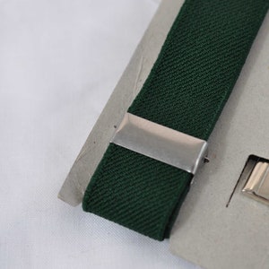 May include: A dark forest green elastic strap featuring a smooth silver-toned metal sliding adjuster and a separate silver-toned metal rectangular clip component, both mounted on a light grey cardboard backing for display and storage purposes.