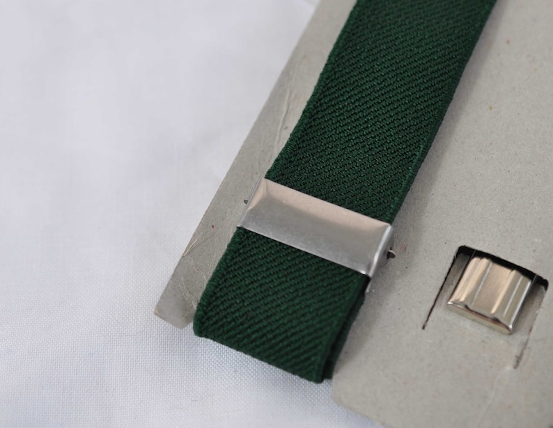 May include: A dark forest green elastic strap featuring a smooth silver-toned metal sliding adjuster and a separate silver-toned metal rectangular clip component, both mounted on a light grey cardboard backing for display and storage purposes.