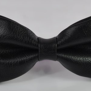 May include: A black bow tie with a textured surface. The bow tie features a classic butterfly shape with a central knot detail. The material has a distinct grain pattern, giving it a matte finish. The edges are folded to create a structured look.