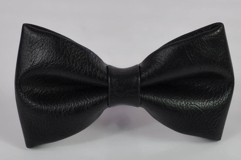 May include: A black bow tie with a textured surface. The bow tie features a classic butterfly shape with a central knot detail. The material has a distinct grain pattern, giving it a matte finish. The edges are folded to create a structured look.
