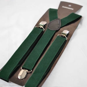 May include: A pair of dark green elastic braces featuring silver-toned metal clips and a brown faux leather triangular back patch. The braces are attached to a brown cardboard display card with the word SUSPENDERS printed at the top.