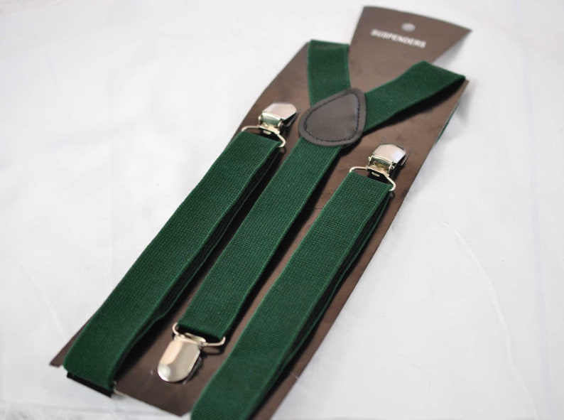 May include: A pair of dark green elastic braces featuring silver-toned metal clips and a brown faux leather triangular back patch. The braces are attached to a brown cardboard display card with the word SUSPENDERS printed at the top.
