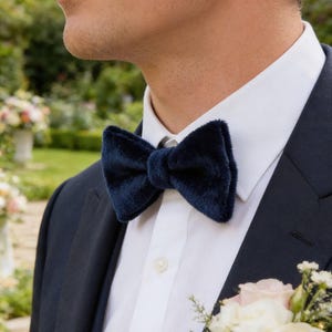 May include: A navy blue velvet bow tie is the focal point of this image. The bow tie is worn with a white dress shirt and a navy blue suit jacket. A small floral arrangement is visible.