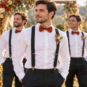 May include: A white long-sleeved button-down shirt paired with black trousers, black braces with gold-toned metal clips, and a solid burnt orange bow tie. A small floral buttonhole with dried flowers is pinned to the left side of the shirt.