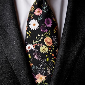 May include: A black necktie featuring a vibrant floral pattern with white, pink, orange, and purple flowers, green leaves, and thin stems. The tie is worn over a crisp white collared shirt and underneath a textured black suit jacket with lapels.