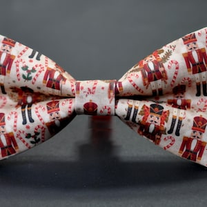 May include: A festive bow tie featuring a repeating pattern of toy soldiers and candy canes on a cream background. The bow tie is a classic shape, perfect for holiday events. The fabric has a slight sheen.