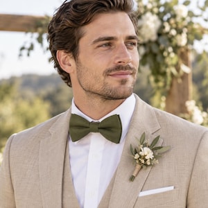 May include: A beige suit jacket worn over a crisp white button-down shirt, featuring a solid olive green bow tie and a small floral boutonniere pinned to the lapel. The boutonniere consists of white flowers, green leaves, and twine wrapping.