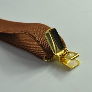 May include: A brown elastic suspender with a gold metal clip. The clip is open and ready to be attached to clothing.