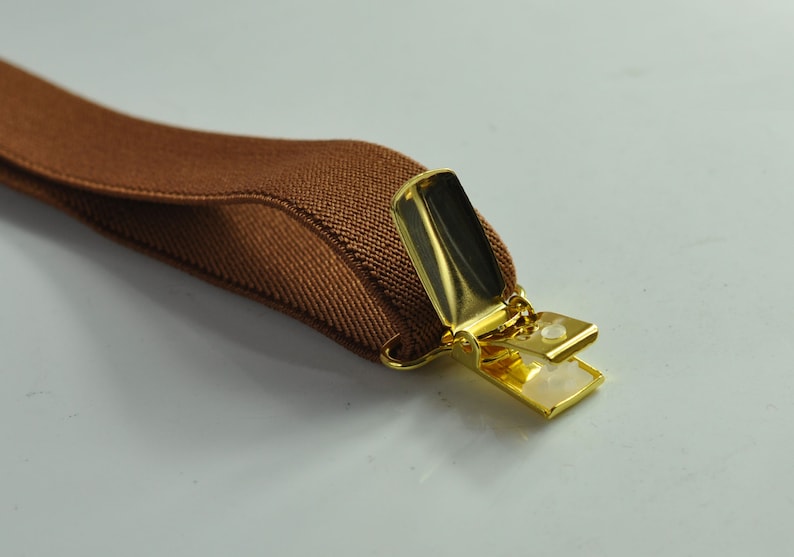 May include: A brown elastic suspender with a gold metal clip. The clip is open and ready to be attached to clothing.