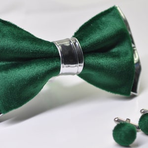 May include: A dark green velvet bow tie with a silver trim and matching cufflinks. The cufflinks are also made of dark green velvet.