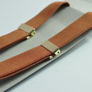 May include: A pair of brown suspenders with gold metal clips. The suspenders are folded in half and laid on a white surface.