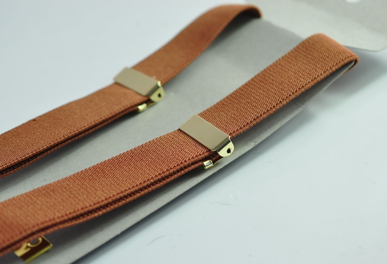 May include: A pair of brown suspenders with gold metal clips. The suspenders are folded in half and laid on a white surface.