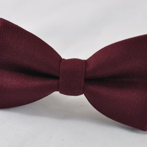 May include: A deep burgundy bow tie featuring a distinct diagonal twill weave texture. The bow tie has a central knot and is attached to a black adjustable strap that extends from both sides, providing a secure fit for formal or casual attire.
