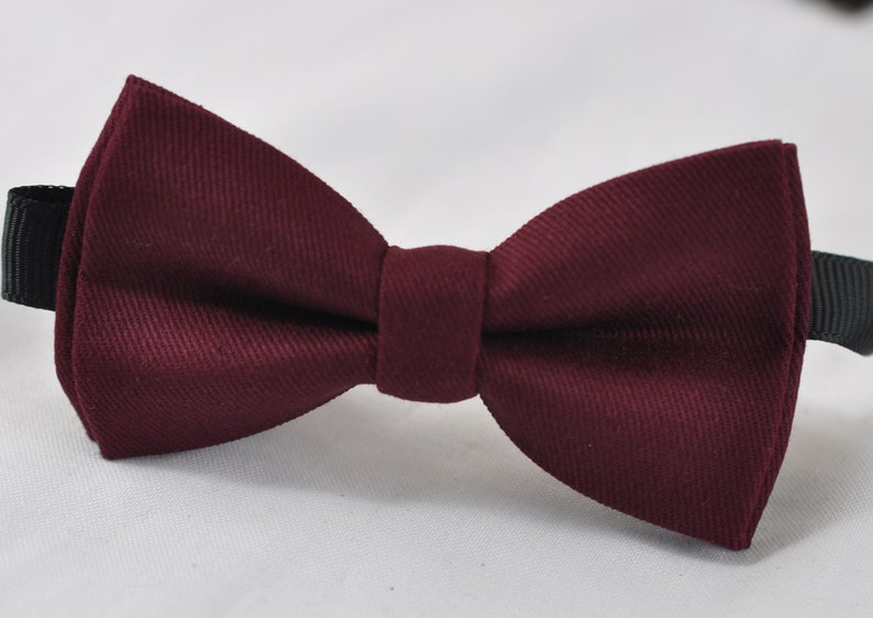 May include: A deep burgundy bow tie featuring a distinct diagonal twill weave texture. The bow tie has a central knot and is attached to a black adjustable strap that extends from both sides, providing a secure fit for formal or casual attire.