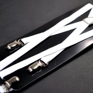 Puede incluir: Tirantes blancos con clips metálicos plateados y un conector triangular. Los tirantes están dispuestos en un patrón entrecruzado sobre un fondo negro. Un accesorio clásico para vestir formal o informal.