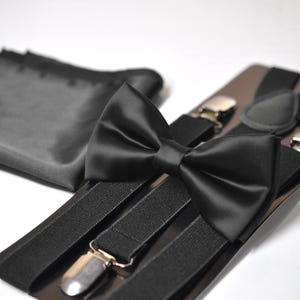 Op de afbeelding: Een zwarte satijnen vlinderdas en bretels set met zilverkleurige metalen clips. Een bijpassende zwarte pochet is ook inbegrepen. De vlinderdas is voorgeknoopt. Deze formele set is ideaal voor bruiloften en feesten.