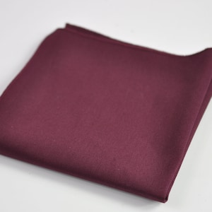 May include: A solid deep burgundy pocket square folded into a neat square shape. The fabric features a smooth, matte texture with clean, finished edges visible along the perimeter of the folded cloth, presenting a uniform and consistent color.