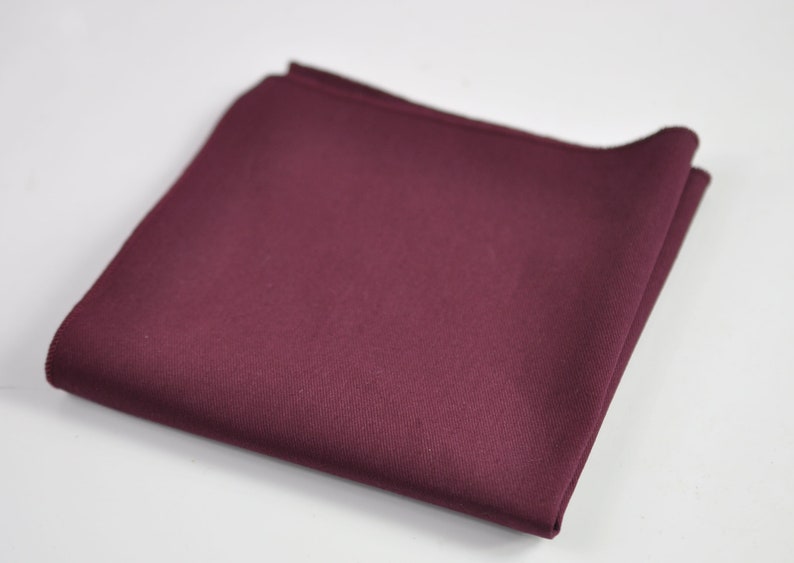 May include: A solid deep burgundy pocket square folded into a neat square shape. The fabric features a smooth, matte texture with clean, finished edges visible along the perimeter of the folded cloth, presenting a uniform and consistent color.