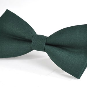 May include: A solid dark forest green bow tie featuring a classic pre-tied shape with a smooth fabric texture and a central knot detail. The accessory includes a thin black adjustable strap extending from the right side of the bow tie structure.