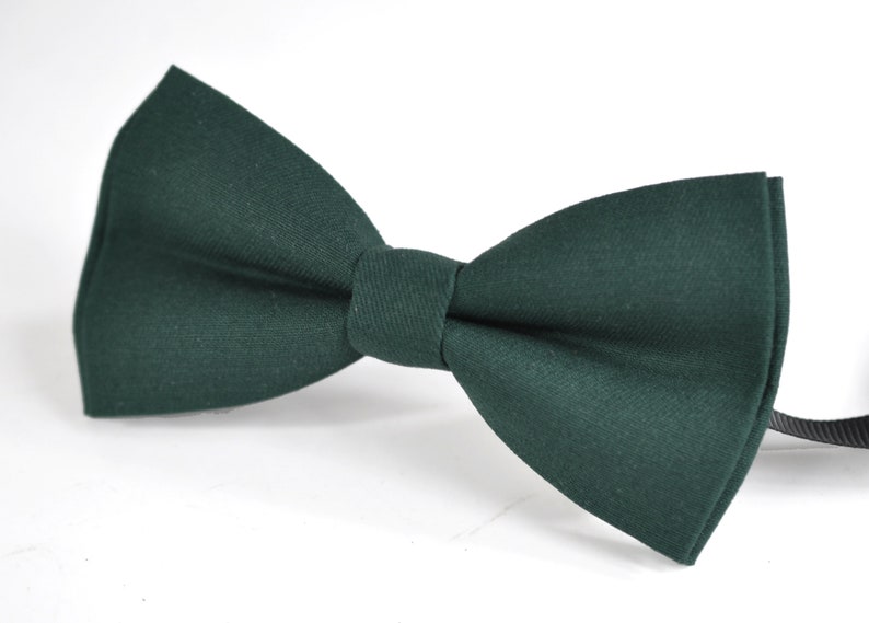 May include: A solid dark forest green bow tie featuring a classic pre-tied shape with a smooth fabric texture and a central knot detail. The accessory includes a thin black adjustable strap extending from the right side of the bow tie structure.