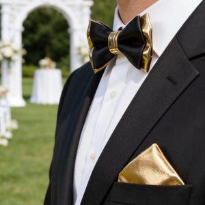 May include: A black tuxedo jacket with a white dress shirt, a black bow tie with gold accents, and a gold pocket square. The bow tie and pocket square add a touch of elegance to the formal attire. The background shows a wedding arch.