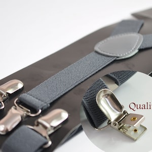 May include: Gray suspenders with silver clips. The suspenders are made of a woven fabric and have a leather patch on the back. The text "Quality clips" is visible in the image.