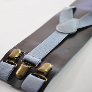 Puede incluir: Tirantes de color azul claro con clips dorados. Los tirantes están empaquetados en una tarjeta negra.