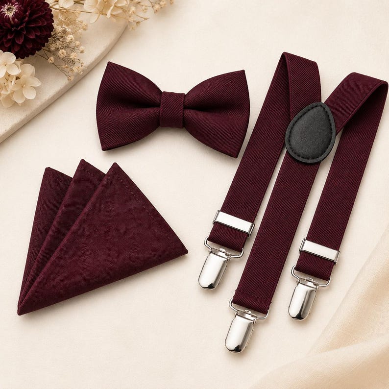 May include: A set consisting of a deep burgundy bow tie, a matching folded pocket square, and adjustable suspenders. The suspenders feature silver-toned metal clips and a black leather-like teardrop-shaped back patch, all in a solid dark red fabric.