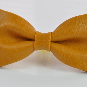 May include: A mustard yellow bow tie with a textured, leather-like surface. The bow tie features a classic pre-tied shape with a central knot detail, showcasing a consistent grain pattern across the entire surface of the accessory.