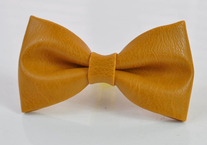 May include: A mustard yellow bow tie with a textured, leather-like surface. The bow tie features a classic pre-tied shape with a central knot detail, showcasing a consistent grain pattern across the entire surface of the accessory.