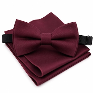 May include: A deep burgundy bow tie with a textured fabric finish, featuring a black adjustable strap with a metal slider. The bow tie is positioned on top of a matching burgundy pocket square, which is folded into a neat, layered square shape.