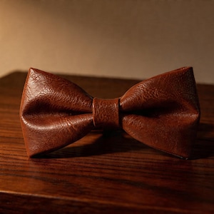 May include: A brown bow tie with a textured, crinkled surface finish. The bow tie features a classic double-loop shape with a central knot detail, showcasing a rich, deep brown color and a distinct, weathered appearance across the entire surface.